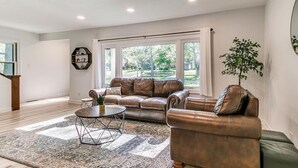 A spacious living room with a leather sofa, a coffee table, and a large window offering a view of the outdoors. - Big Groups~Game Room~5Bedroom~Kids Toys~Office (Des Moines)