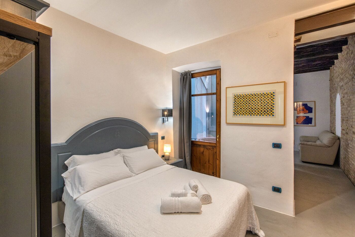 Un dormitorio con una cama, una mesita de noche, una ventana con cortinas, una puerta y un cuadro colgado en la pared.