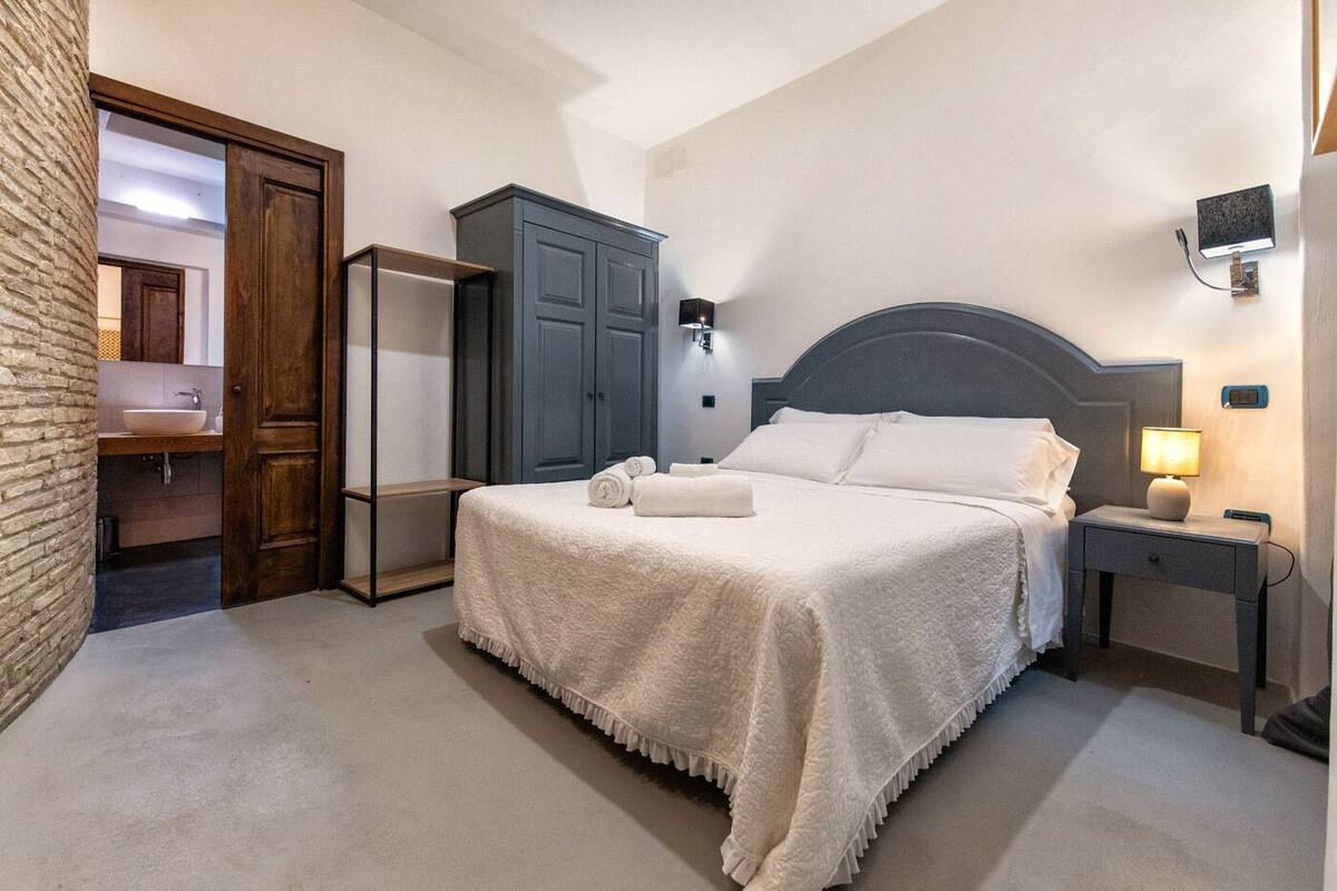 Een slaapkamer met een bed, nachtkastje, lamp, kledingkast en een zichtbare badkamer door een open deur.