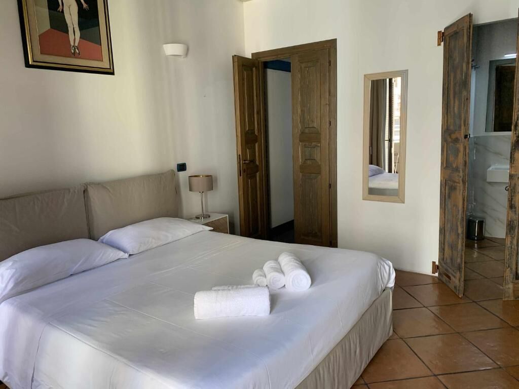 Una habitación de hotel con cama, mesita de noche, lámpara, espejo y una puerta que da a otra estancia.