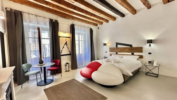 A modern hotel room with exposed wooden beams, a large bed with white bedding, a red and white bean bag chair, a small round table with a chair, and a large window with sheer curtains.