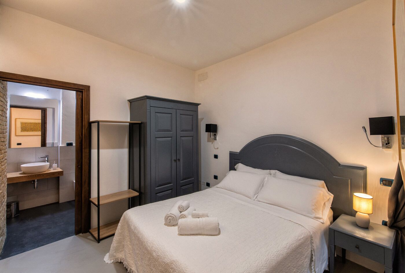 A hotel room with a bed, a wardrobe, a bathroom with a sink, and a nightstand with a lamp.