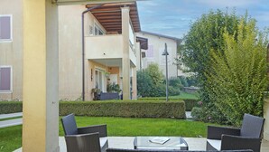 An outdoor patio area with wicker furniture, a glass-top table, and a view of a two-story building with balconies and a garden. - Le Corti Caterina C3 by Wonderful Italy (Lonato del Garda)