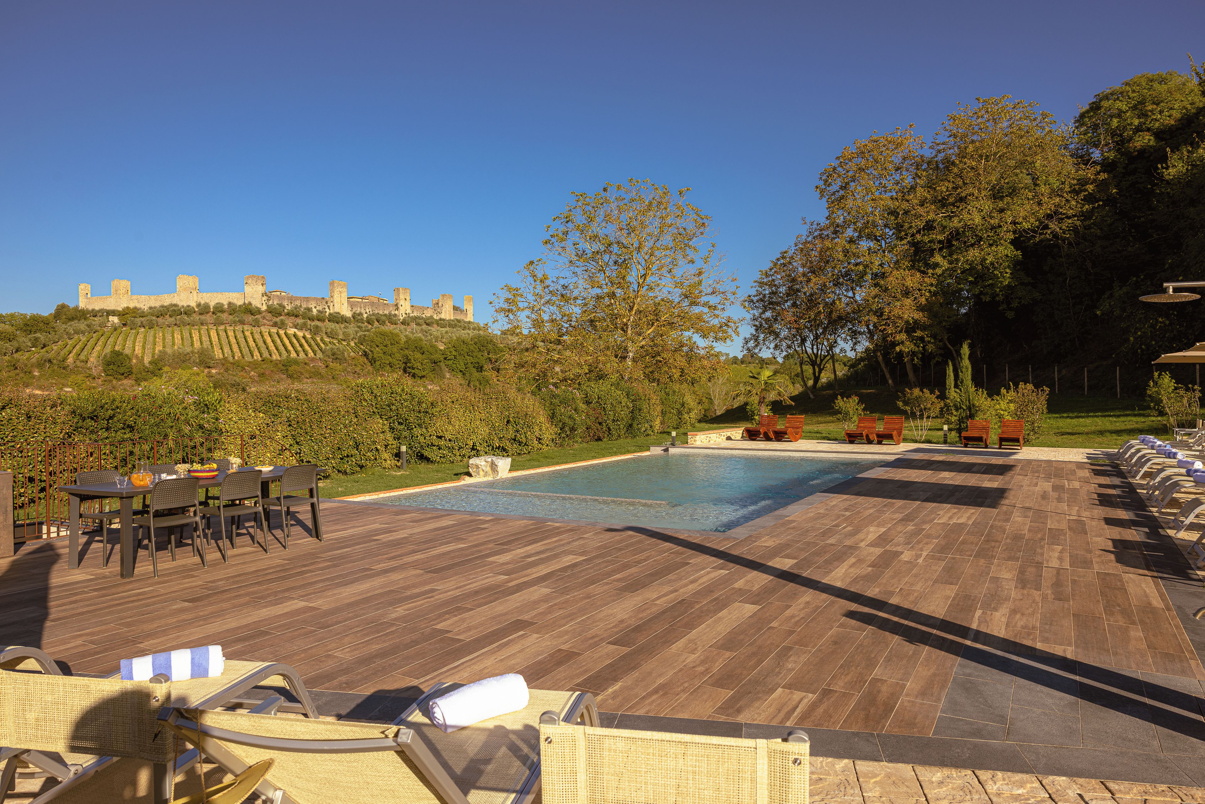 A pool area with lounge chairs and a table, a view of a castle on a hill, and a well-maintained garden.
