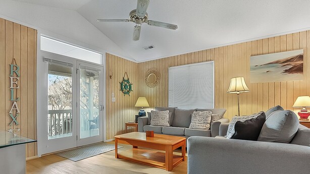 A living room with a wooden floor, a grey sofa, a wooden coffee table, a ceiling fan, and a window with blinds.