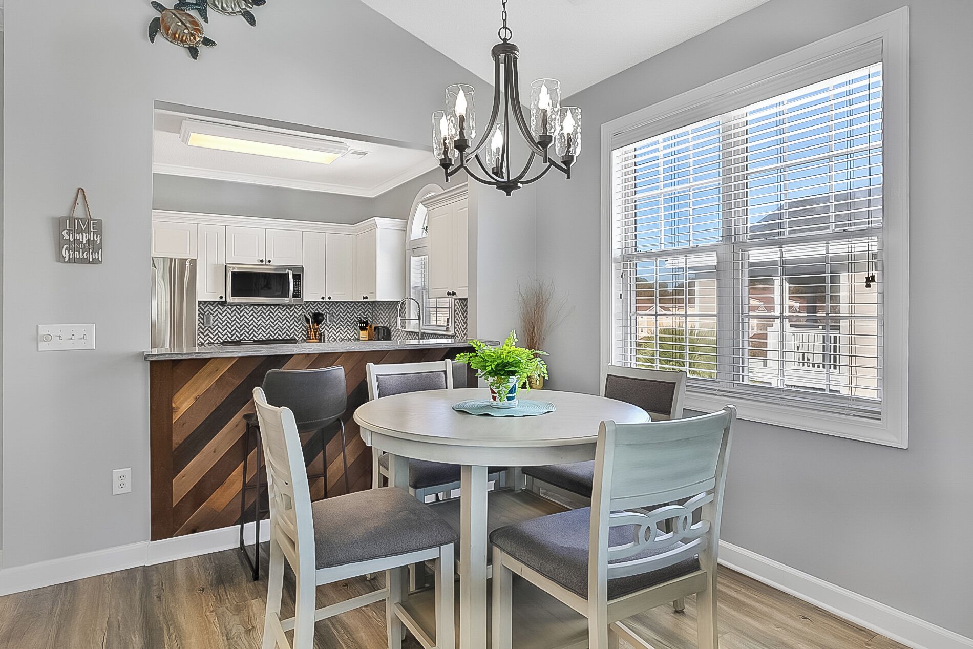A modern kitchen with white cabinets, a microwave, and a chandelier. There is a dining table with chairs and a potted plant.