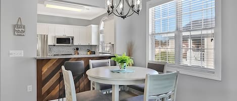 A modern kitchen with white cabinets, a microwave, and a chandelier. There is a dining table with chairs and a potted plant.