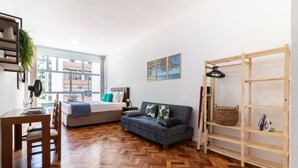 A modern living room with a sofa, a dining table with chairs, a wooden herringbone floor, a large window, and a wooden shelving unit. - Pi502 Zona4 in Rio de Janeiro (Rio de Janeiro)