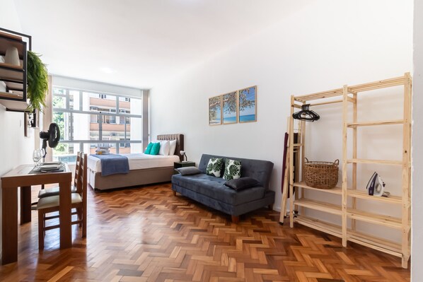 Una sala de estar moderna con un sofá, una mesa de comedor con sillas, piso de madera con patrón de espiga, un ventanal grande y una estantería de madera.
