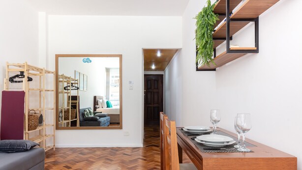 A modern living room with a wooden herringbone floor, a grey sofa, a wooden dining table with chairs, a mirror reflecting a cozy living area, and a hanging plant. - Pi502 Zona4 in Rio de Janeiro (Rio de Janeiro)