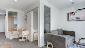 Un salon moderne avec un canapé gris, une petite table ronde et une table basse en verre. On y trouve également un coin repas avec une table et des chaises blanches, et une cuisine visible à travers une porte ouverte.