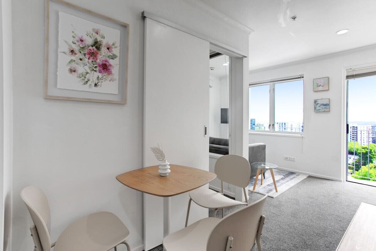 A modern room with a wooden table and chairs, a framed floral artwork, and a view of the city through large windows.