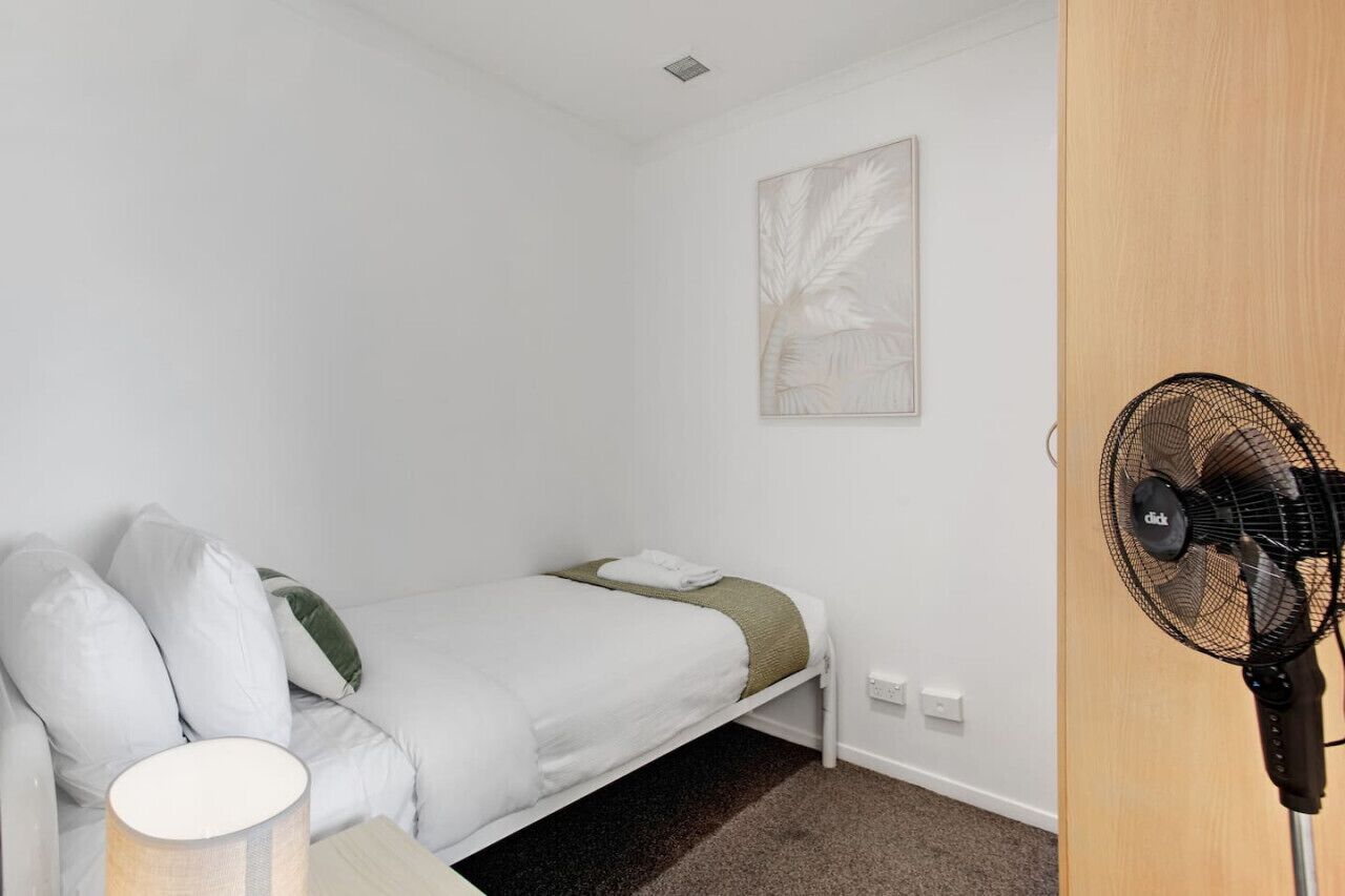 A small, neatly arranged bedroom with a single bed, a bedside table with a lamp, a wall-mounted fan, and a framed artwork above the bed.
