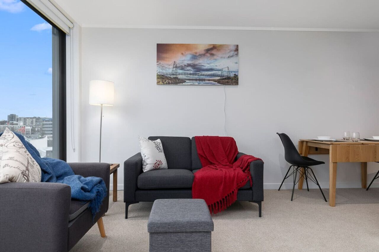 Un salon moderne avec un canapé gris, un plaid rouge, un pouf gris et une table à manger avec des chaises. Un tableau représentant un pont et un coucher de soleil est accroché au mur.