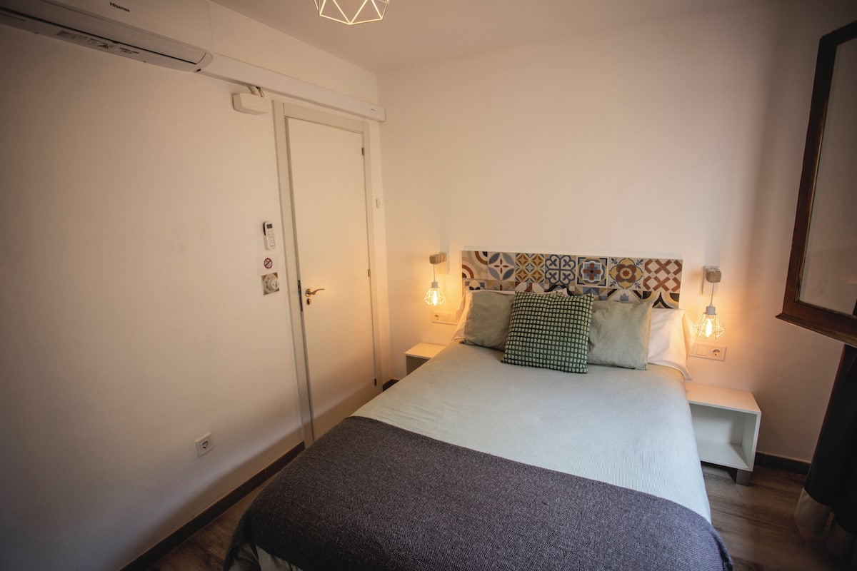 A bedroom with a bed, two bedside tables, a nightstand, a wall-mounted light fixture, and a door.