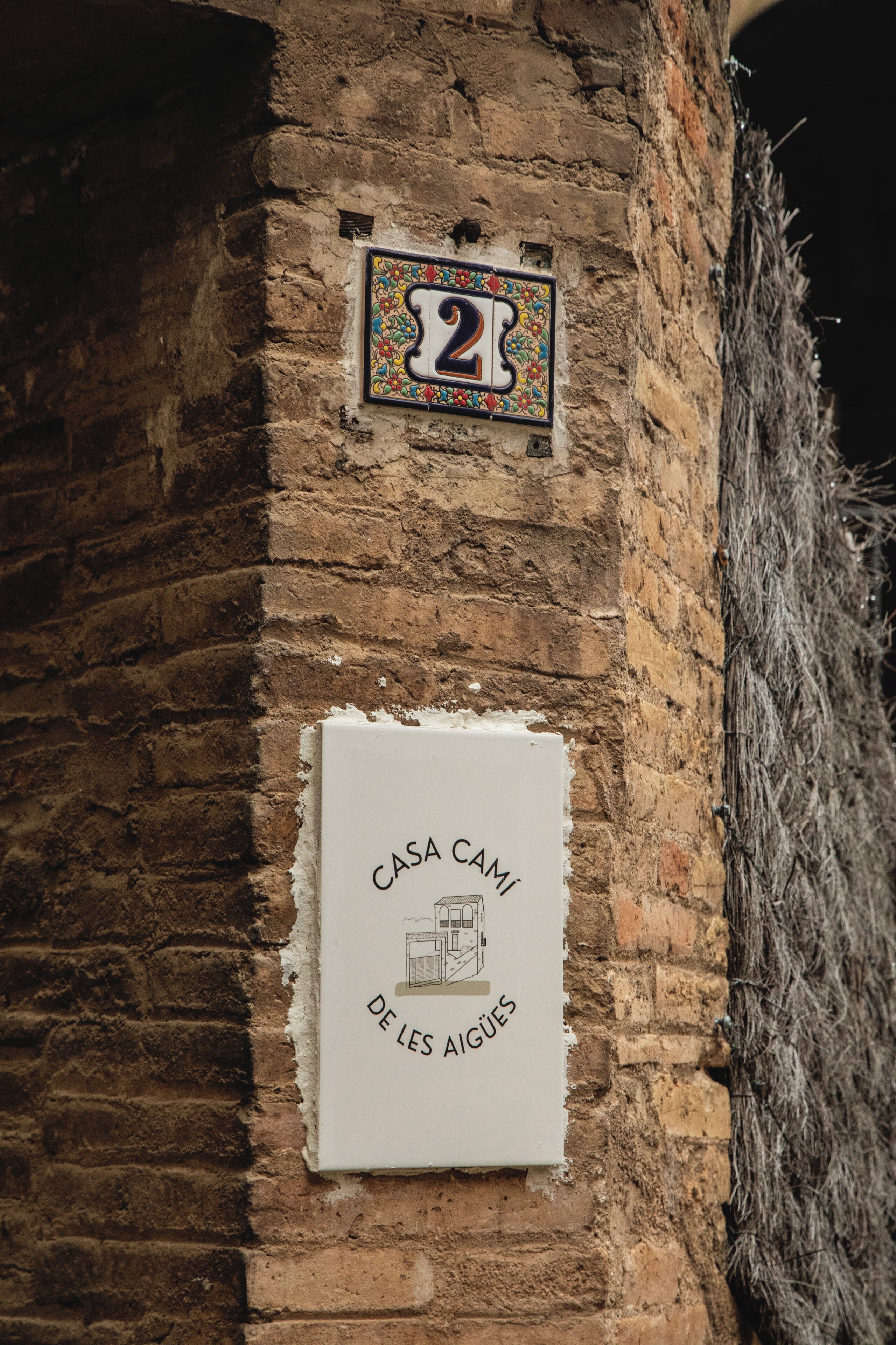 Ein Gebäude mit einer Ziegelwand und zwei Schildern: eines mit der Zahl '2' und das andere mit 'CASA CAMI DE LES AIGUES'.