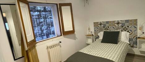 A bedroom with a bed, a window with curtains, a radiator, an air conditioning unit, and a hanging plant.