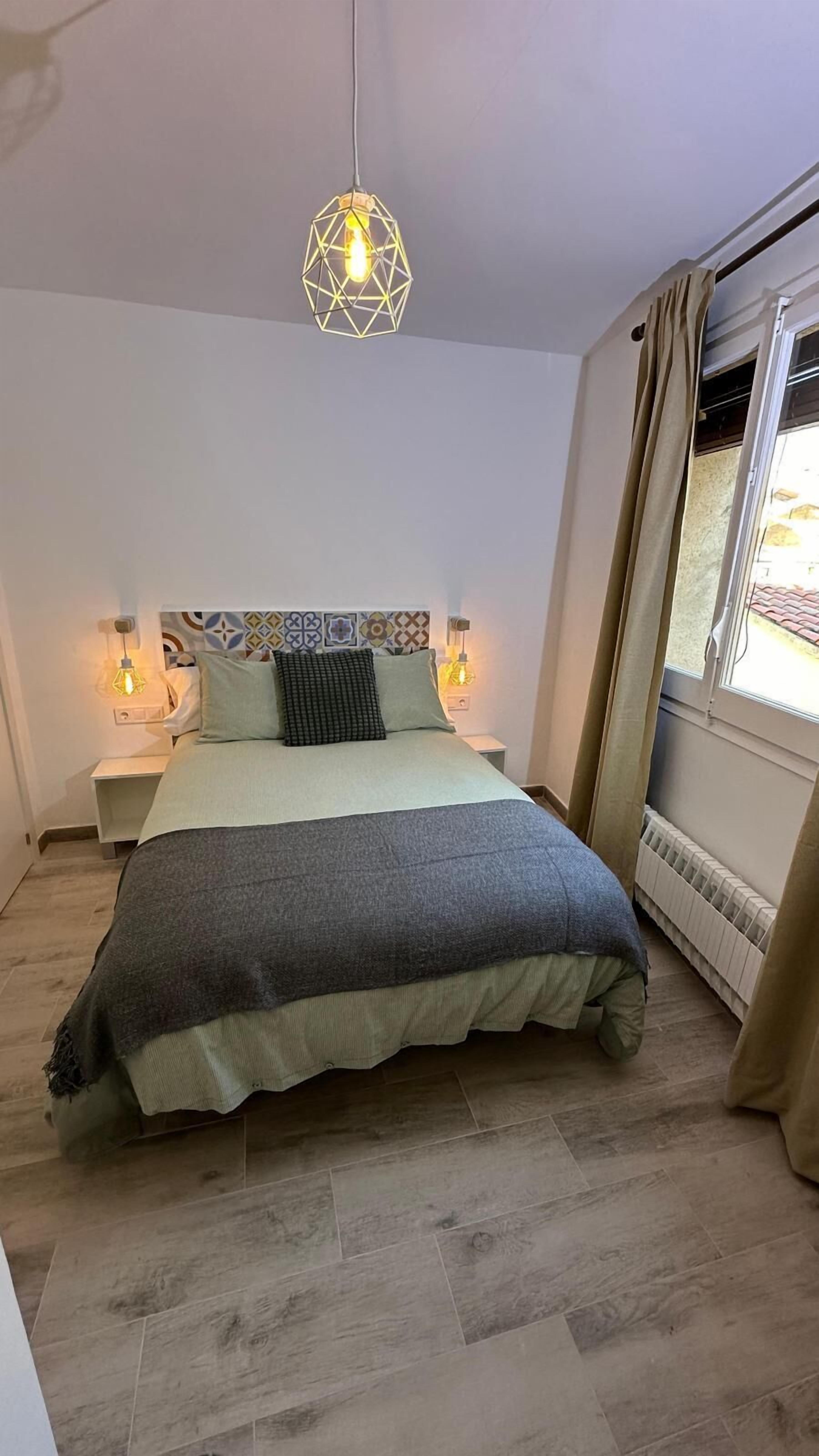 A bedroom with a bed, two bedside tables, a geometric pendant light, a radiator, and a window with curtains.