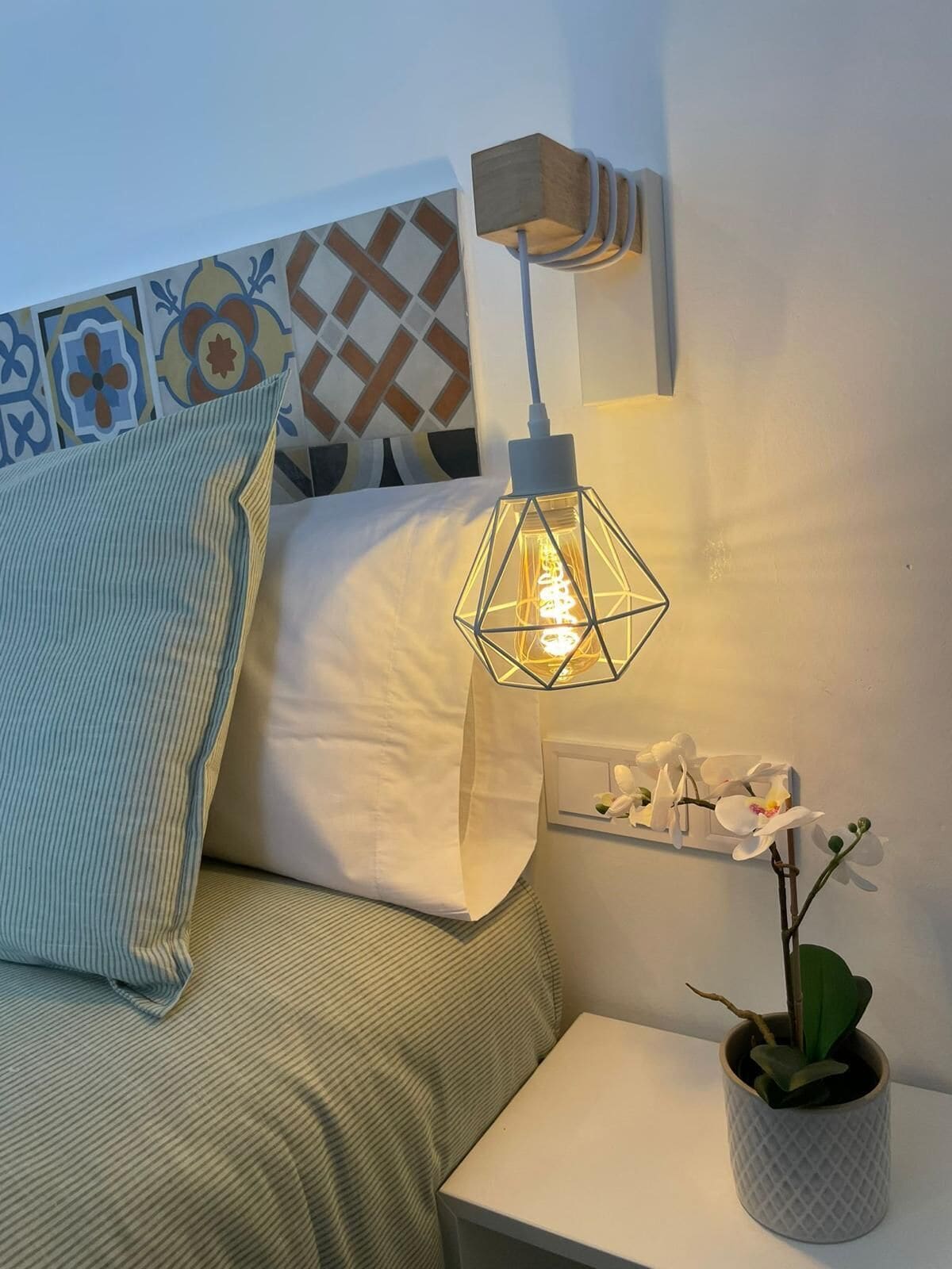 A neatly arranged bedroom with a bed, a bedside table with a potted plant, a wall-mounted light fixture, and a decorative wall panel.