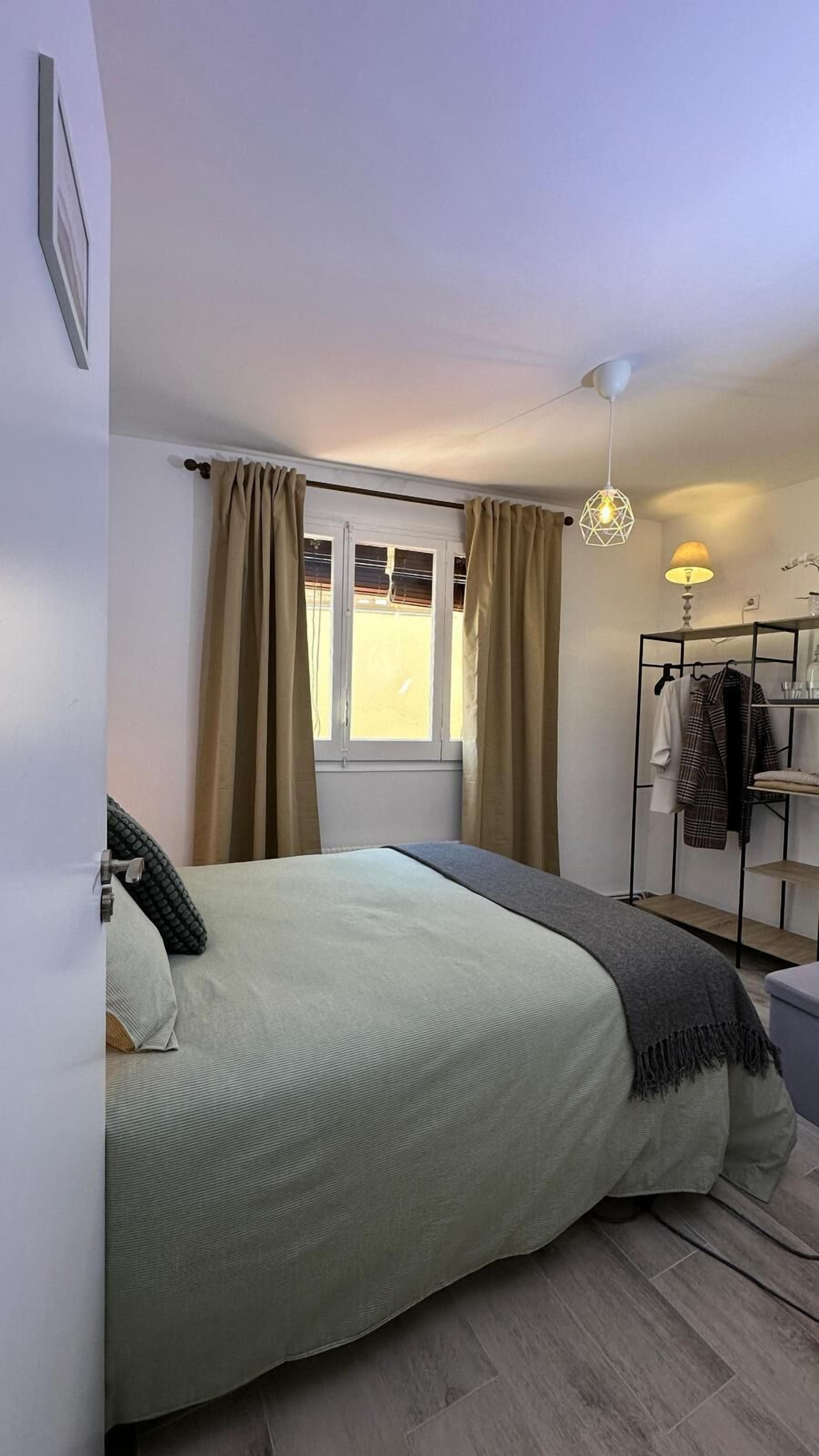 Un dormitorio con cama, ventana con cortinas, mesita de noche, lámpara y perchero.