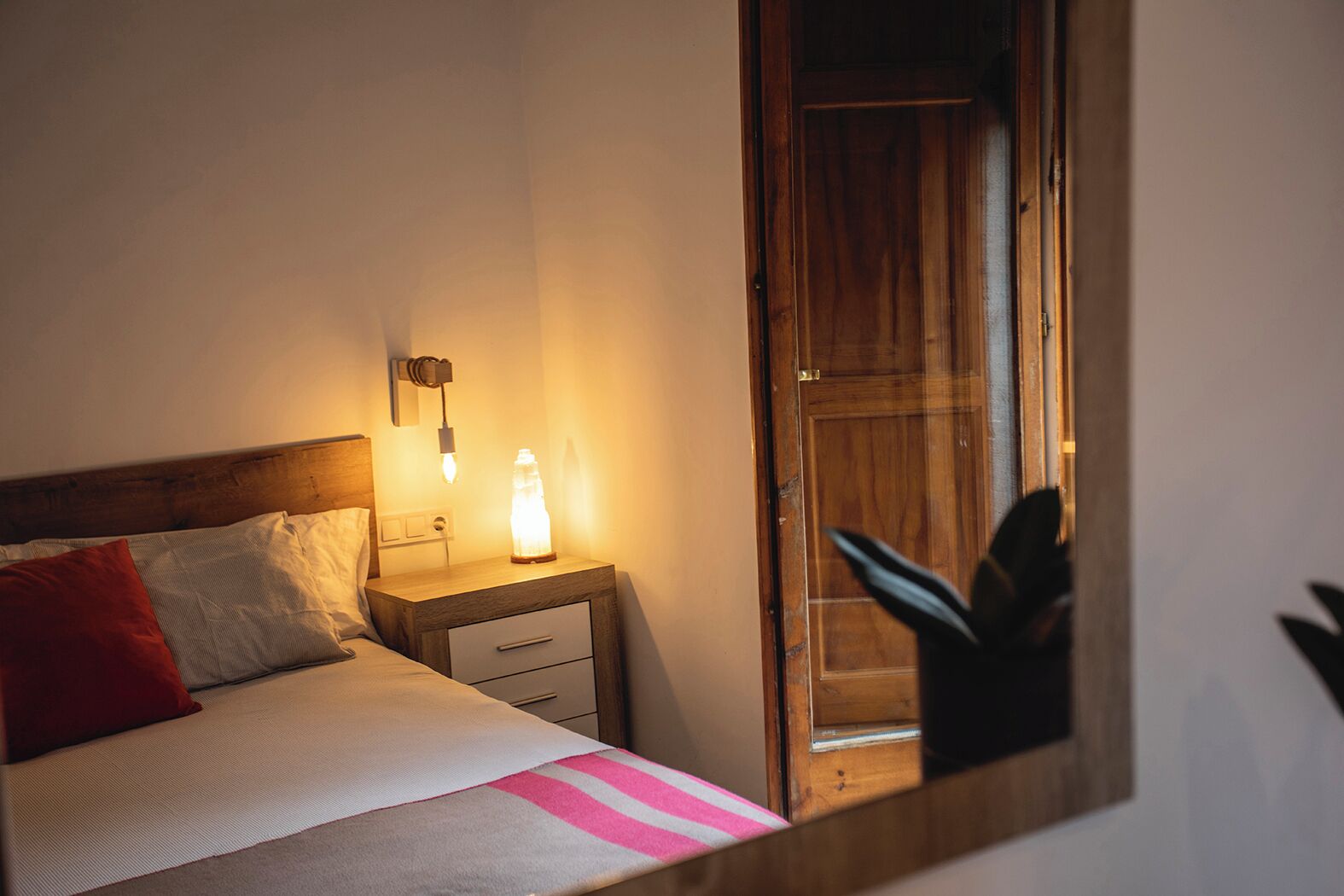 Une chambre à coucher avec un lit, une table de chevet, une commode et un miroir qui reflète une porte en bois.