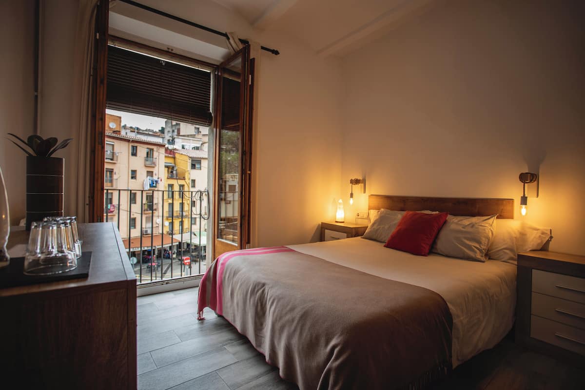 A bedroom with a large bed, bedside tables, a nightstand, a window with a view of buildings, and a balcony.
