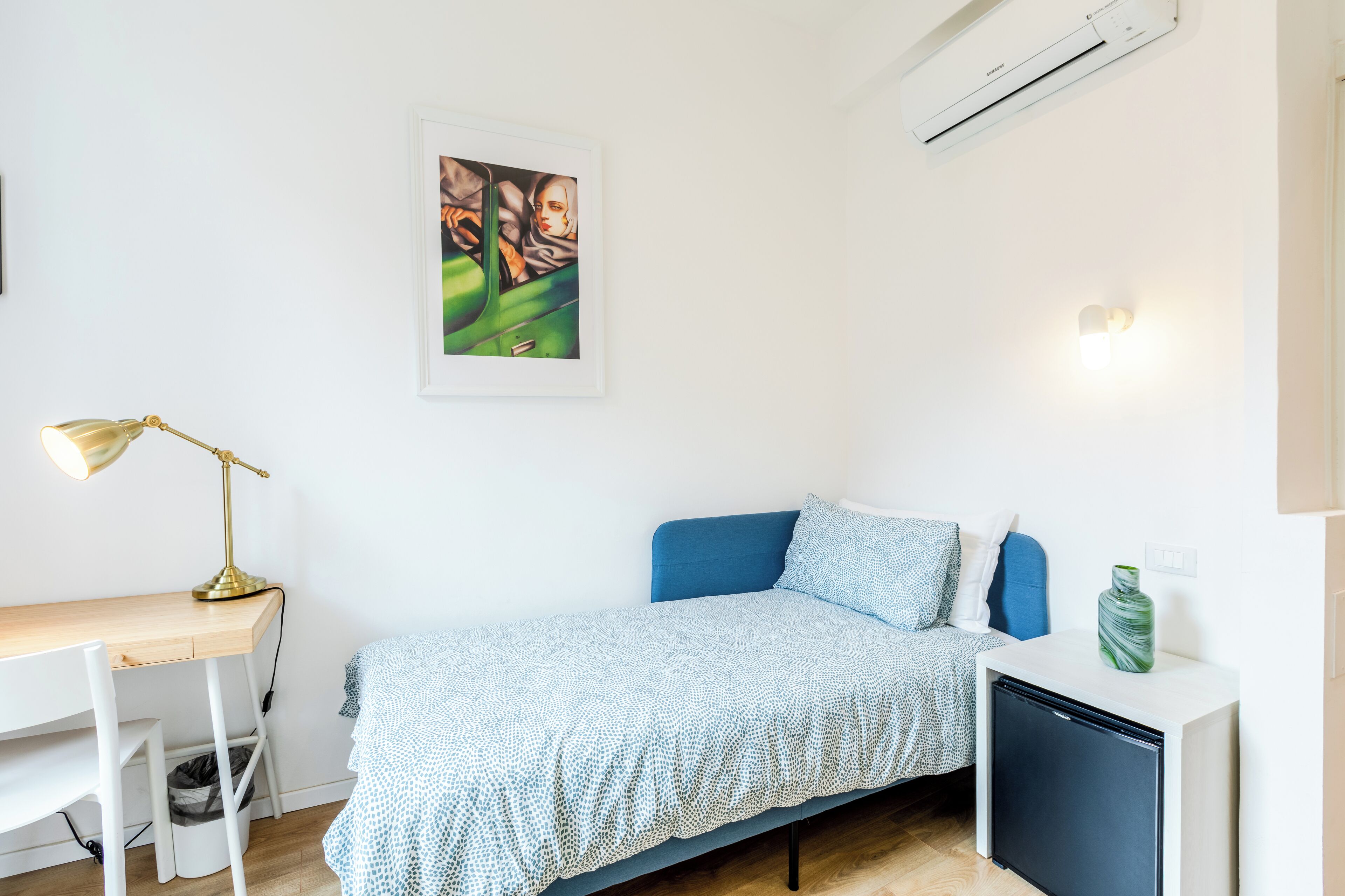 A small, neatly arranged bedroom with a bed, desk, and a small refrigerator.