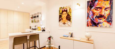 Uma cozinha moderna com armários brancos, um espaço de bar e dois banquetes. Há duas pinturas na parede: uma retrata uma mulher com gatos e a outra é um abstrato colorido.