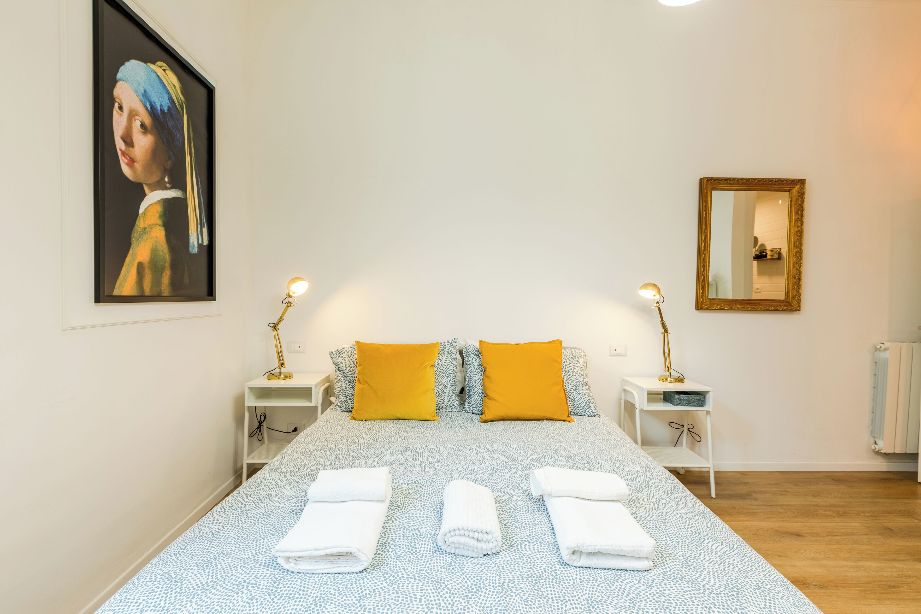 Een netjes opgemaakt bed met twee kussens, een nachtkastje met een lamp, een ingelijste foto aan de muur, en een spiegel.