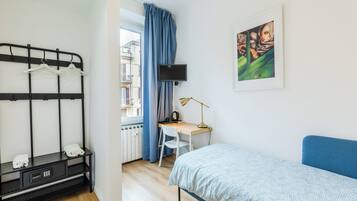 A bedroom with a bunk bed, a desk with a chair, a bed with a blue headboard, a TV on the wall, and a window with blue curtains.