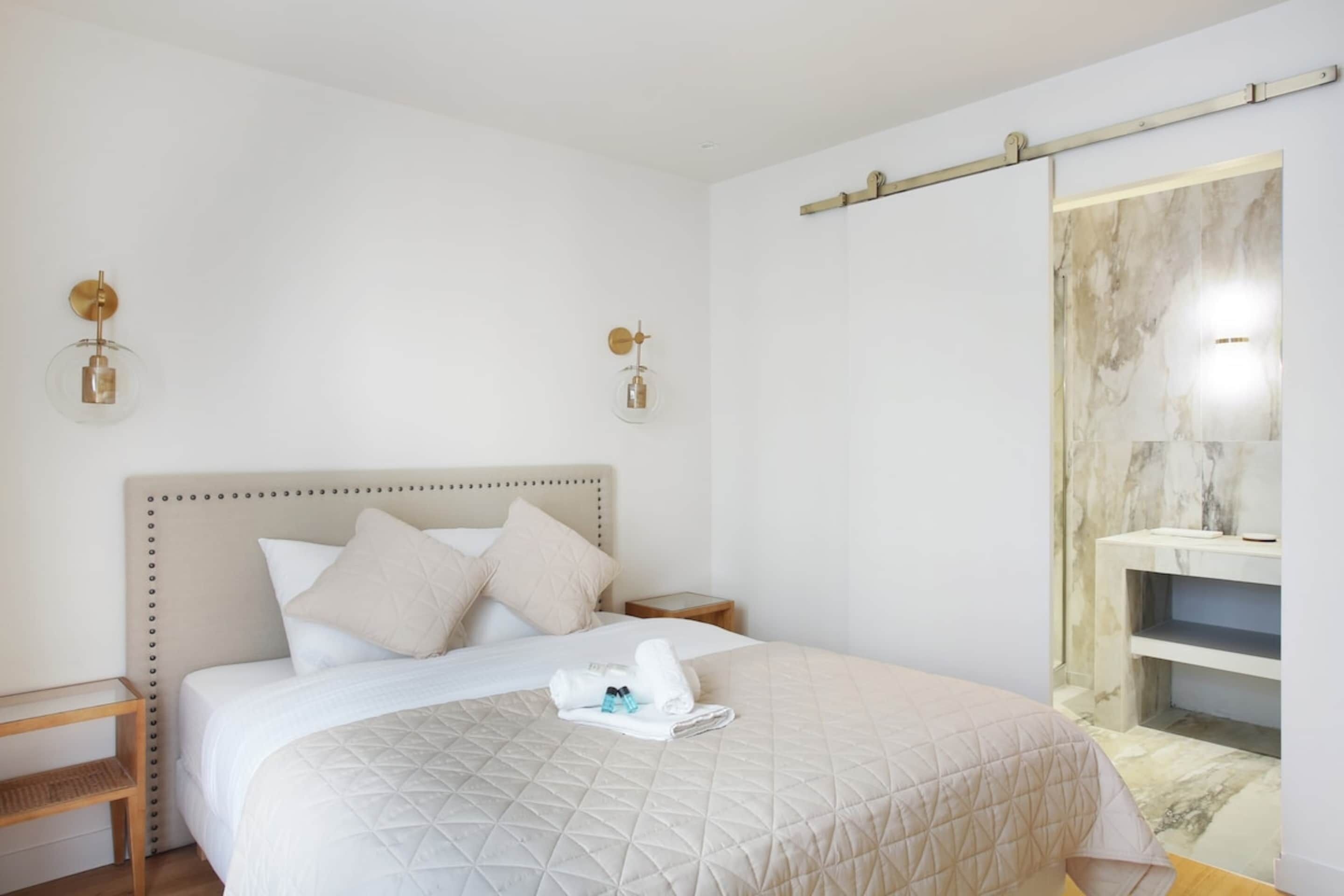 A hotel room with a bed, bedside tables, a nightstand, a wall-mounted light fixture, and a marble-patterned wall.