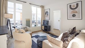 A modern living room with a sofa, armchair, and a coffee table. There is a television on a stand, a lamp, and artwork on the wall.