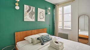 A hotel room with a green wall, a wooden headboard, two artworks, a bed with white linens, a folded towel, and a bedside table with a lamp.
