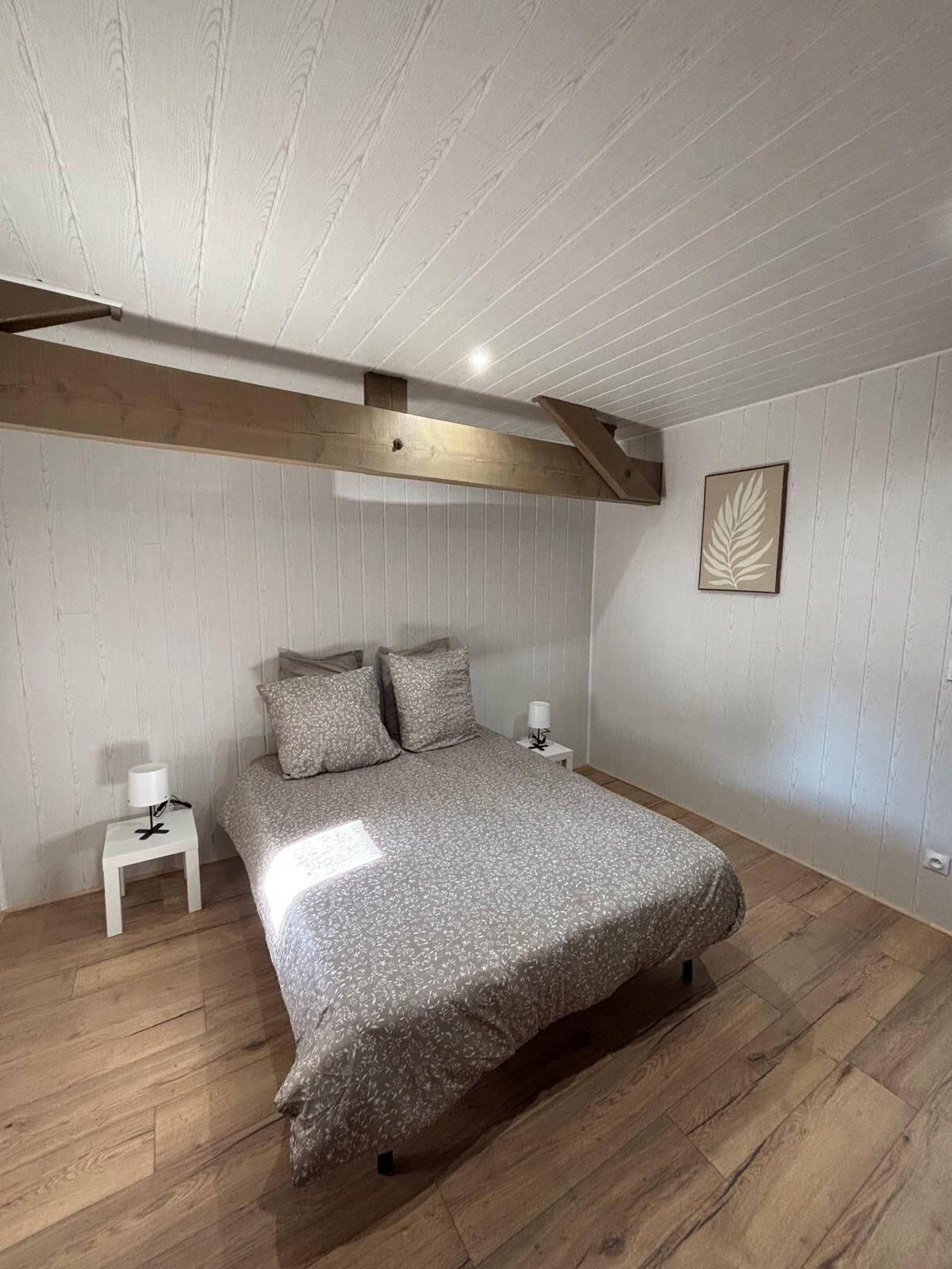 Een slaapkamer met een houten plafond, een bed met een grijze geborduurde spreideken, twee nachtkastjes met lampen, en een wanddecoratie met een bladmotief.