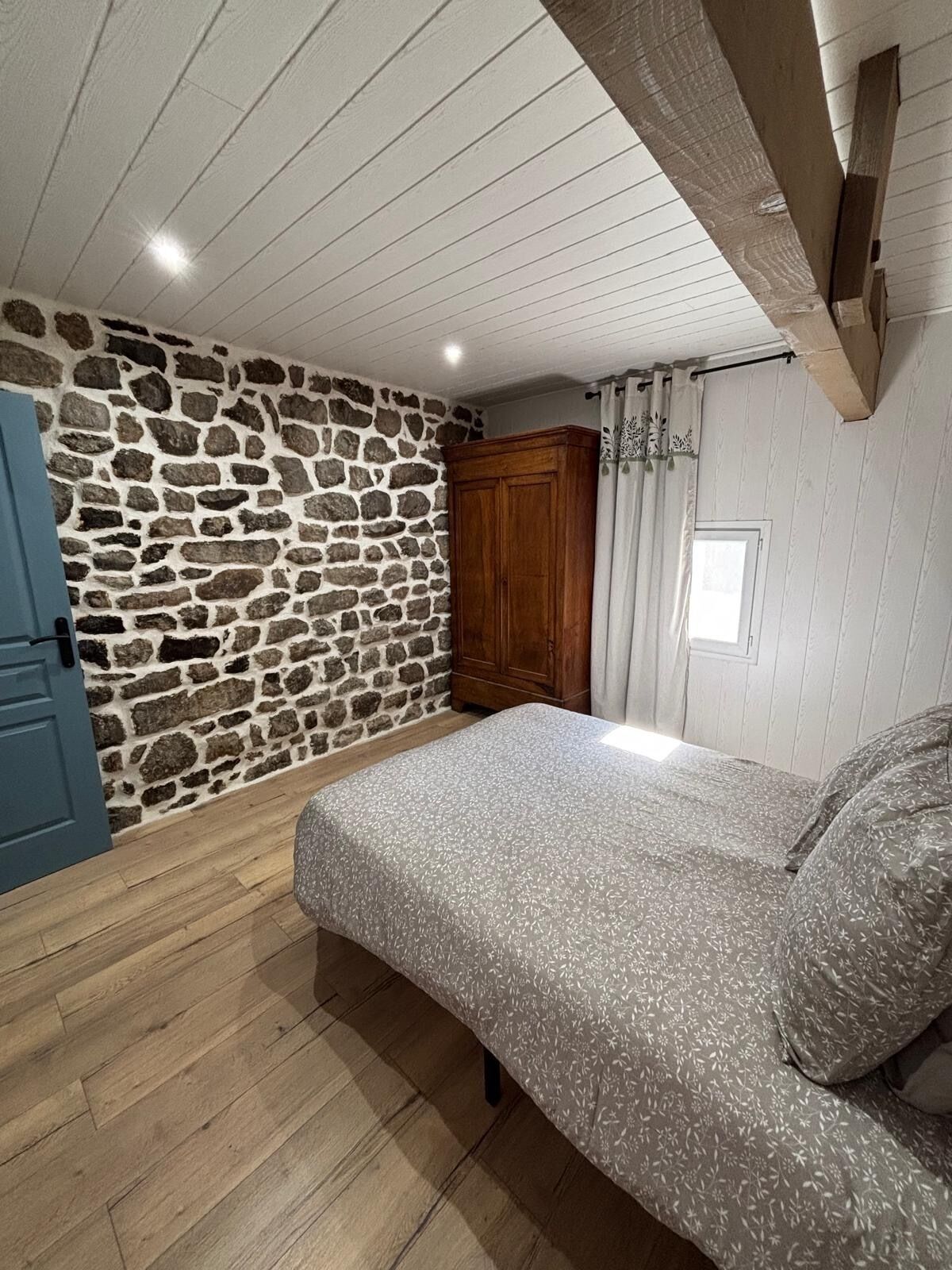 A bedroom with a stone wall, wooden ceiling beams, a bed with patterned bedding, and a wardrobe.