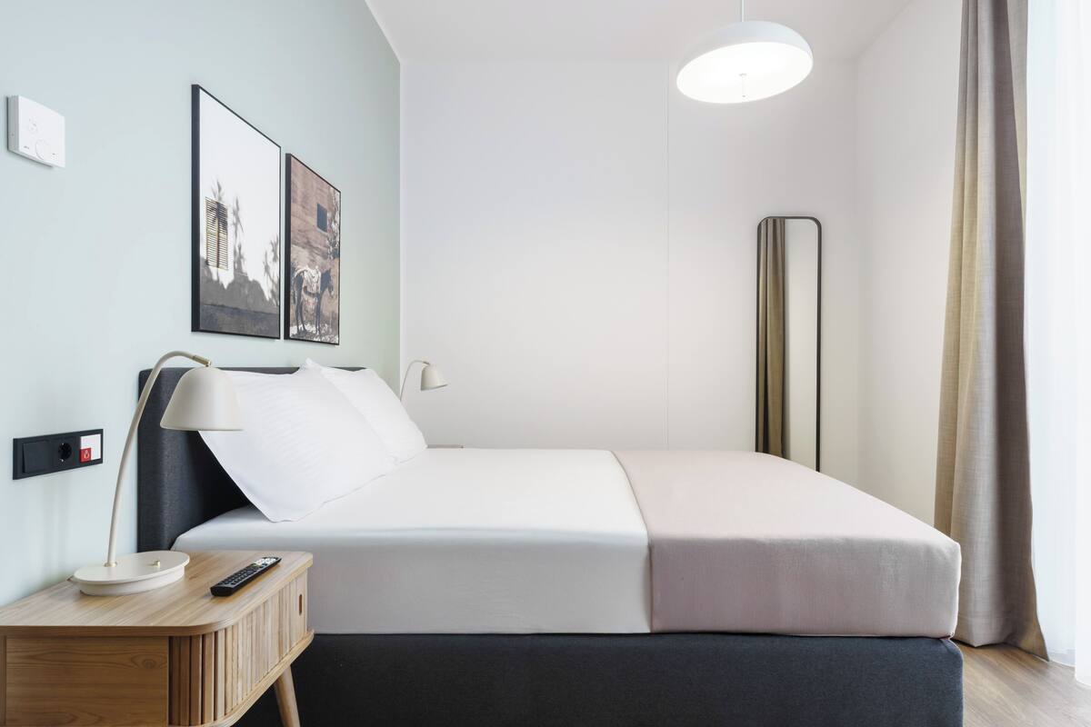 A modern hotel room with a neatly made bed, a bedside table with a lamp, and a wall-mounted control panel.