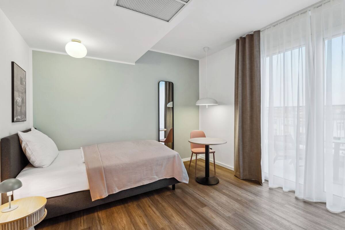 A modern hotel room with a bed, bedside table, chair, and a view of the city through sheer curtains.