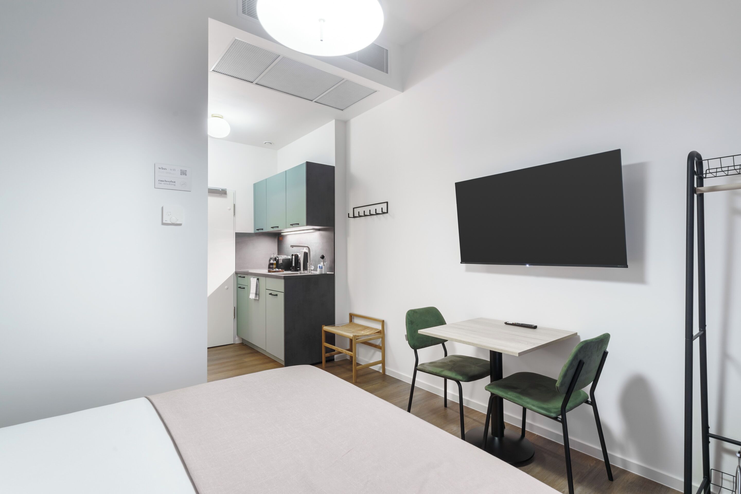 A modern hotel room with a bed, a small dining table with chairs, a kitchenette, and a TV mounted on the wall.