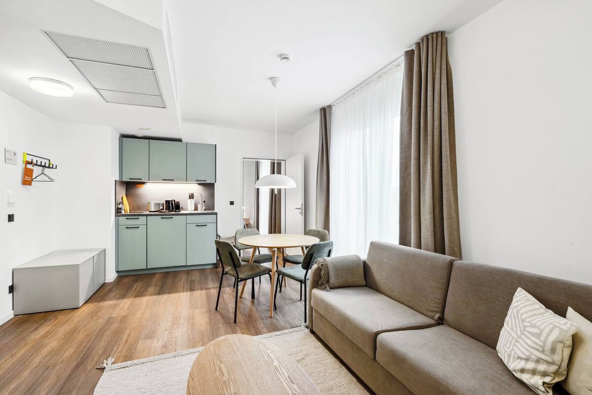 A modern living room with a sofa, a dining table with chairs, a kitchen area, and a large window with curtains.