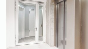 A hallway with a glass door leading to a room, a wall-mounted elevator, and a concrete wall. - Limehome Berlin Sybelstraße (Berlin)