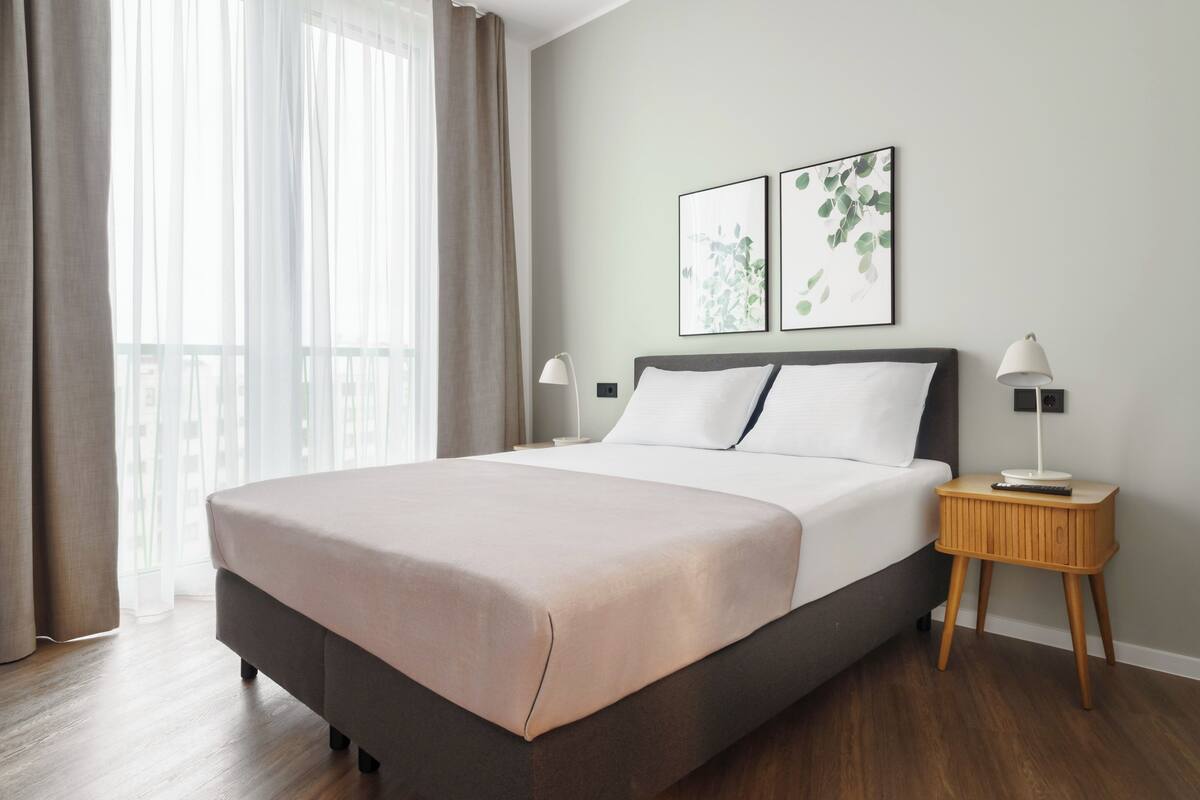 A modern bedroom with a large bed, a bedside table with a lamp, and two framed botanical prints on the wall.