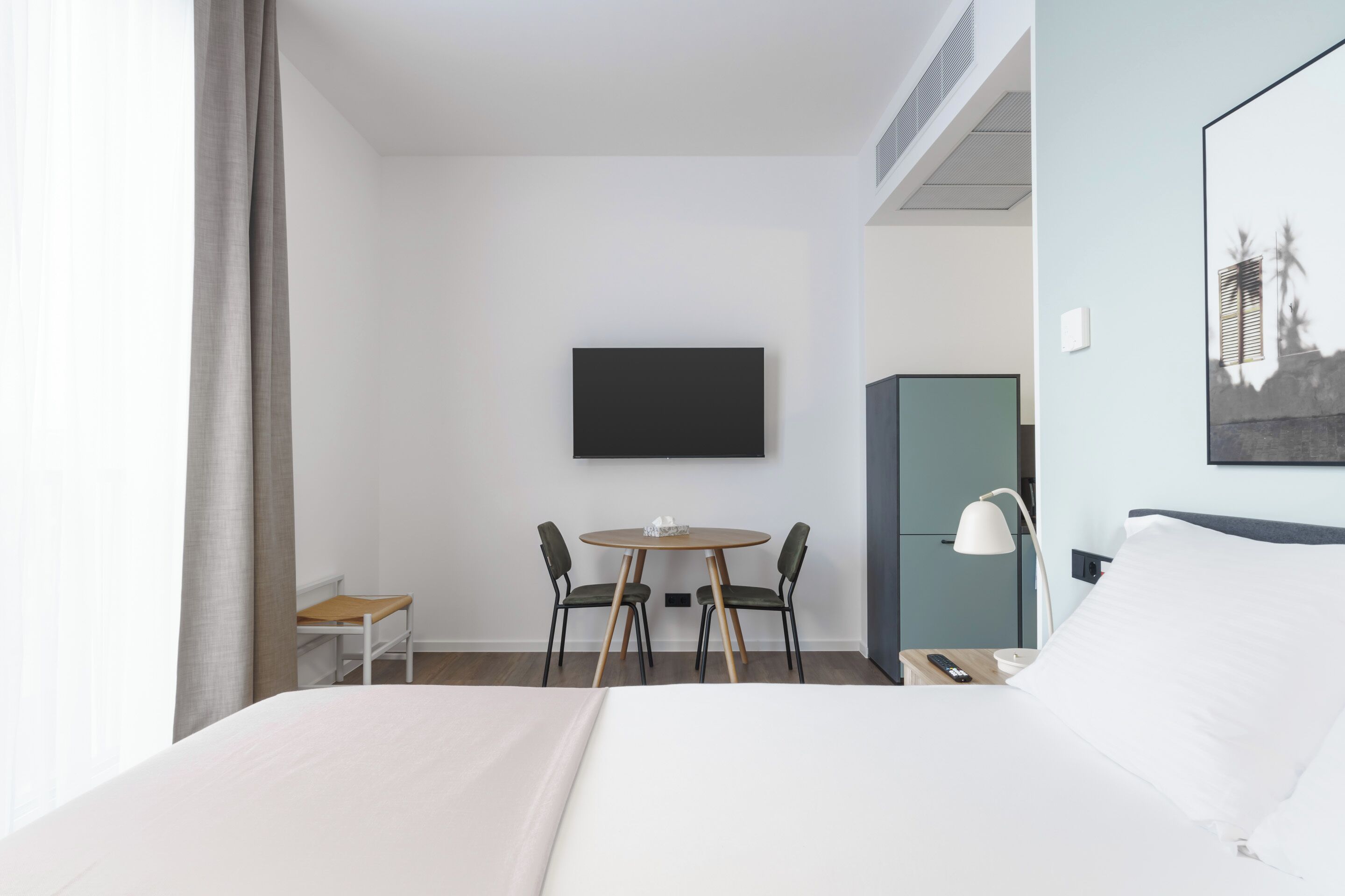 A hotel room with a bed, a desk with a chair, a television mounted on the wall, and a nightstand with a lamp.
