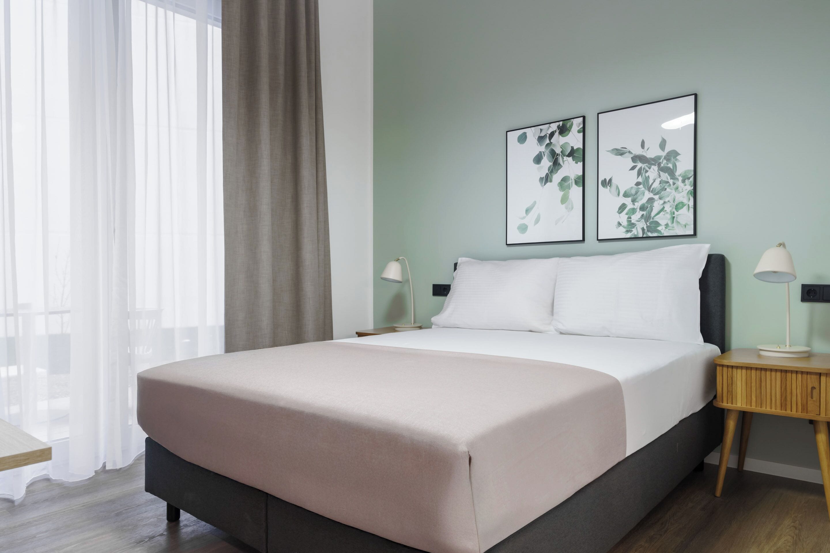 A neatly made bed with a light-colored bedspread, two bedside tables with lamps, and two framed botanical prints on the wall.