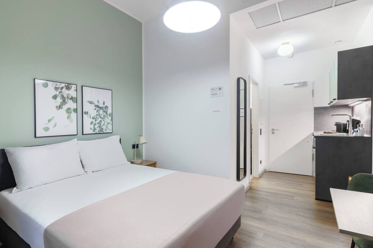A hotel room with a bed, a nightstand, a desk, and a kitchenette.