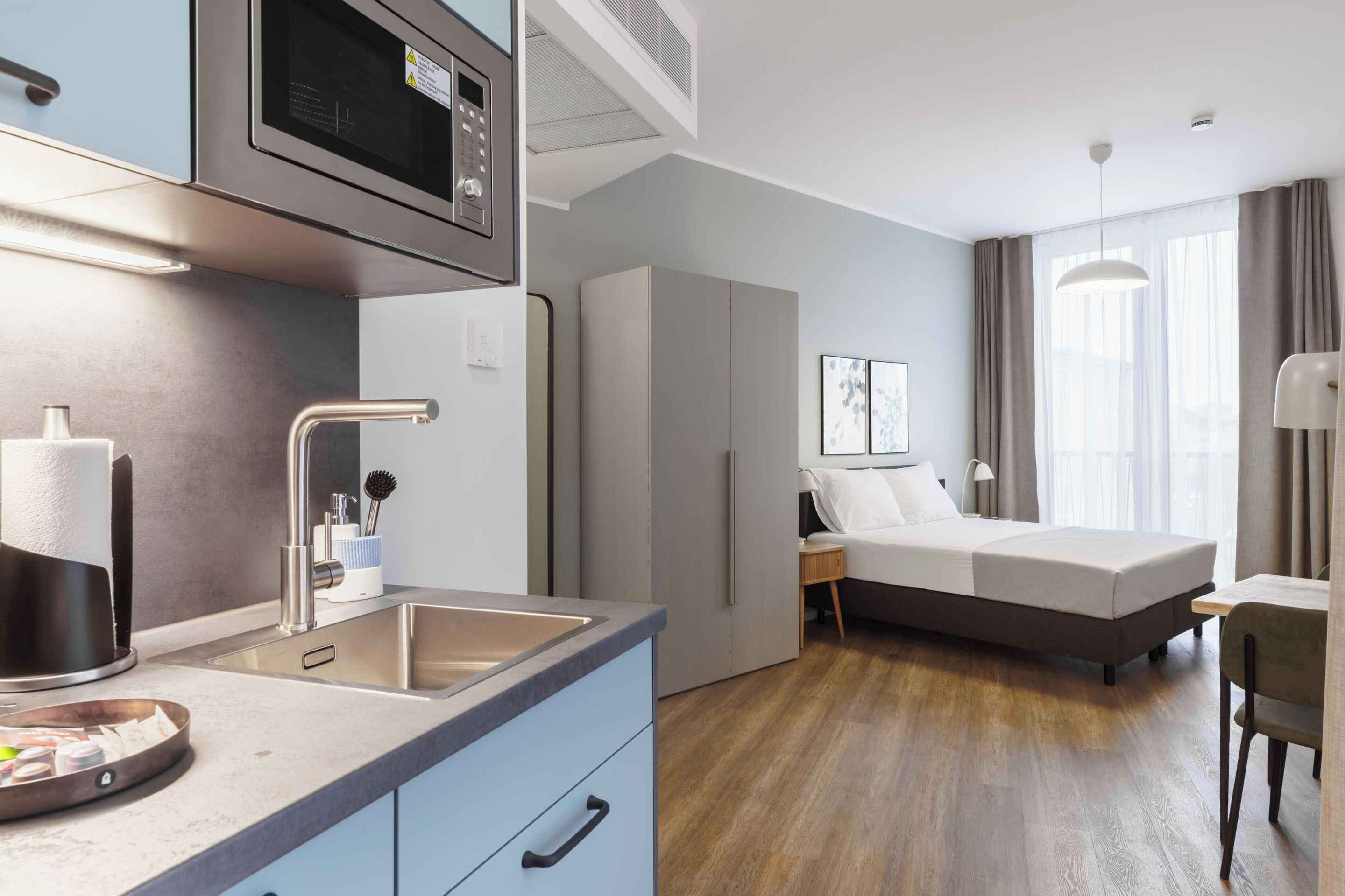 Ein modernes Hotelzimmer mit Küchenzeile, einem Bett, einem Schreibtisch und einem großen Fenster.