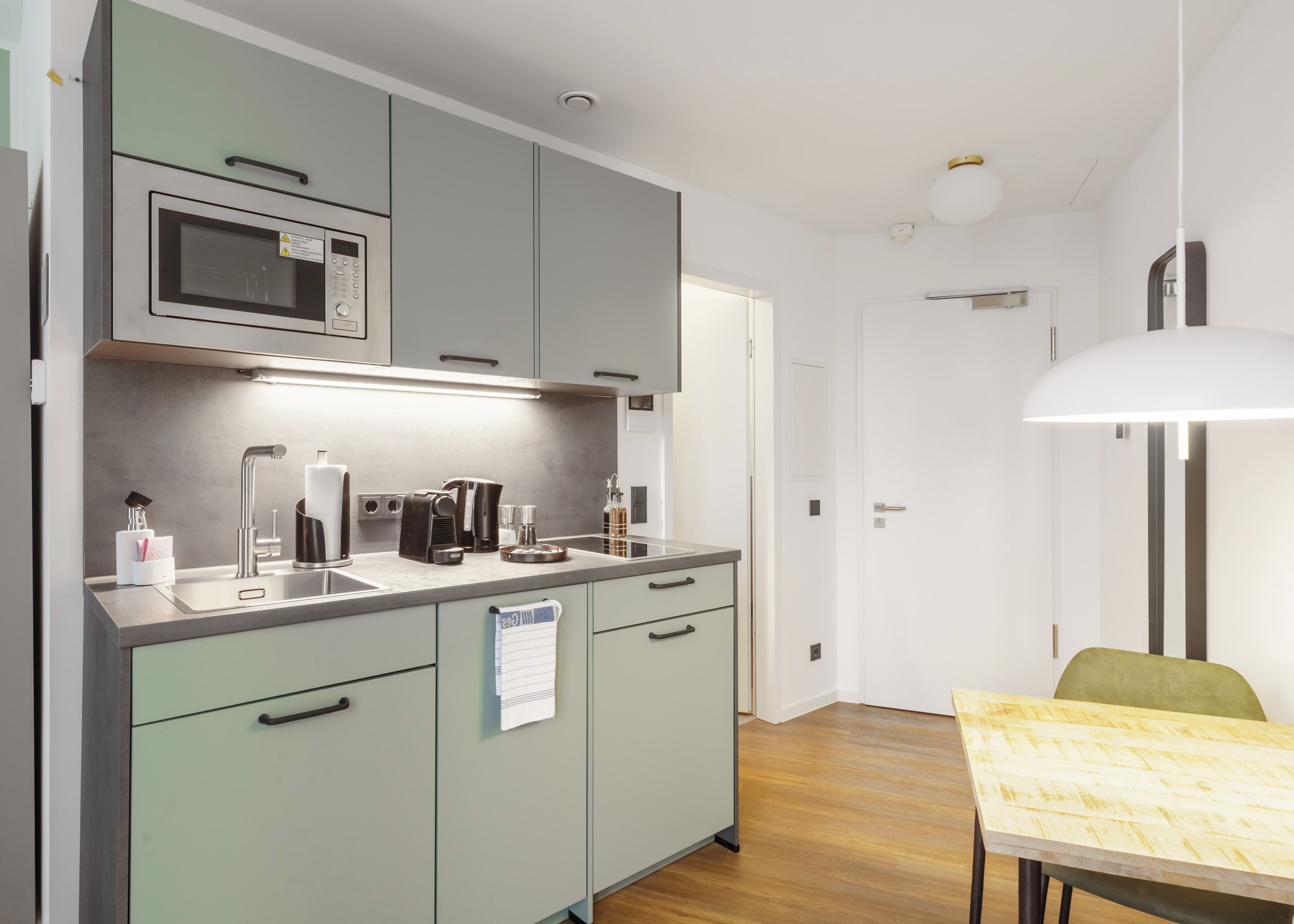 Une cuisine compacte équipée d’un four à micro-ondes, d’une machine à café et d’une bouilloire. Elle comprend un évier avec un robinet, ainsi qu’une petite table avec une chaise. Les meubles de la cuisine sont de couleur vert clair et le plan de travail est blanc.