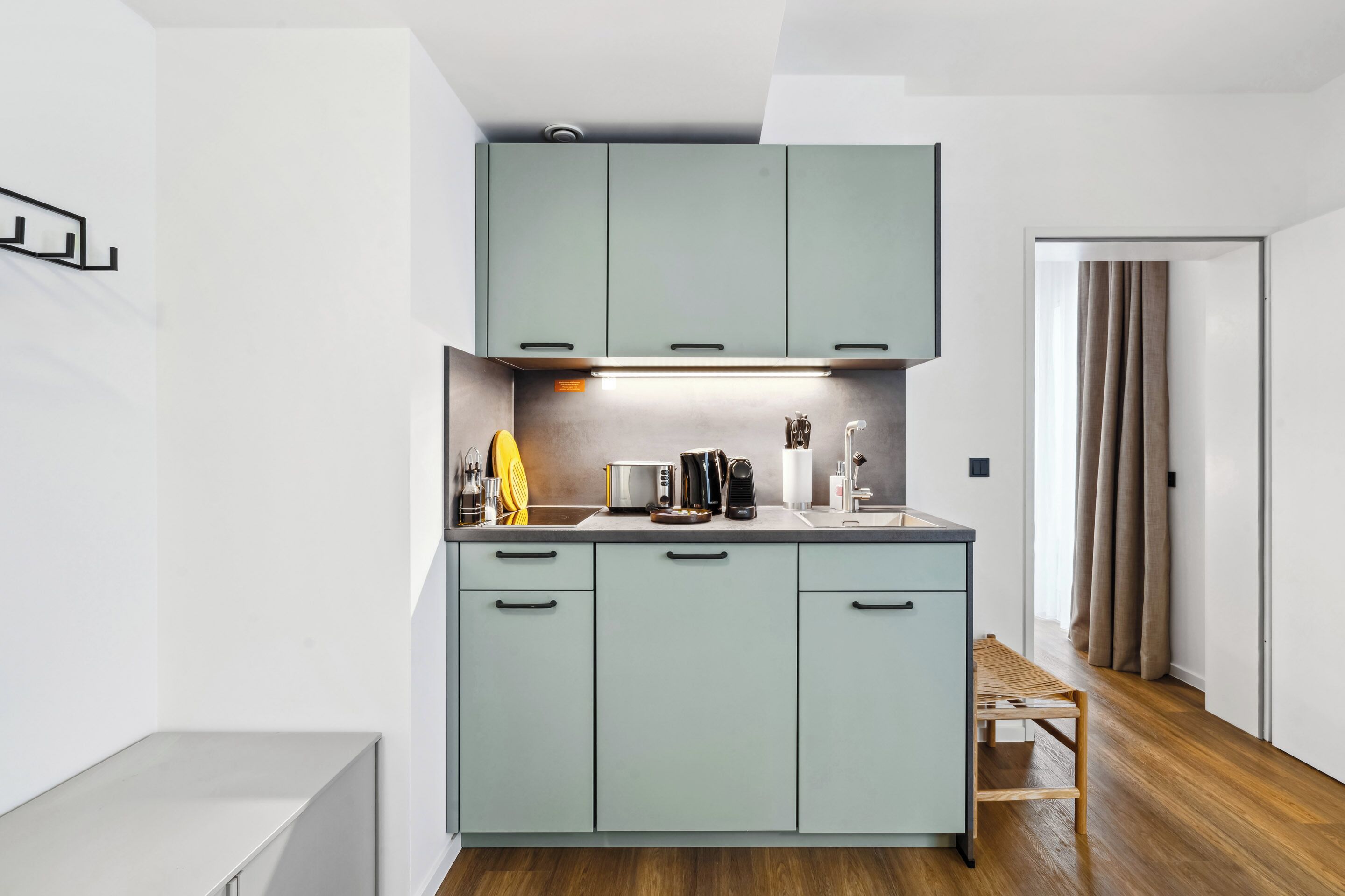 Eine moderne Küche mit hellgrünen Schränken, einer Arbeitsplatte mit Wasserkocher, Toaster und weiteren Geräten. Es steht eine hölzerne Bank dort und eine Tür führt in einen anderen Raum.