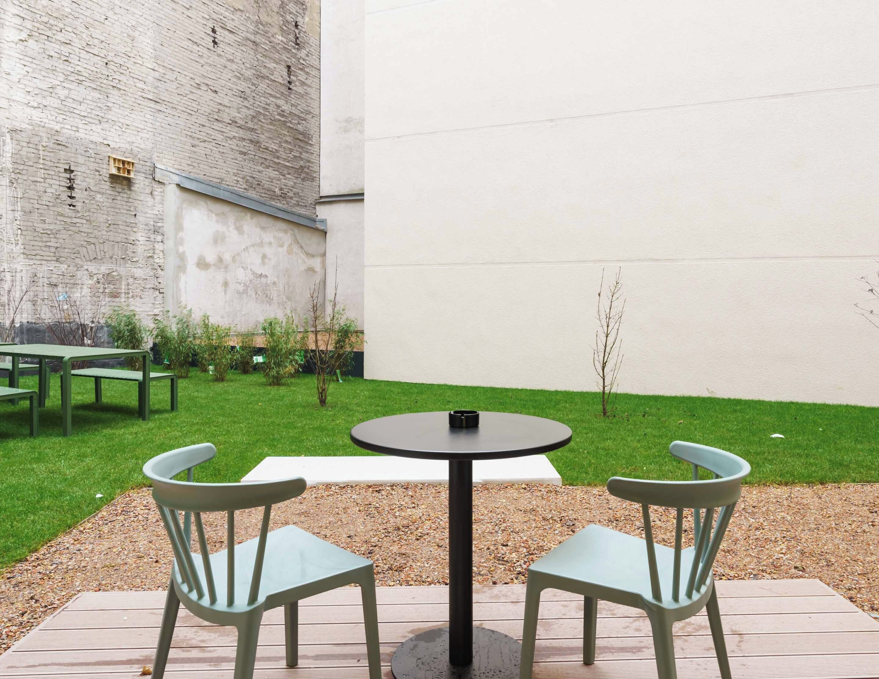 A patio area with a round table and two chairs, a small garden with grass and plants, and a brick wall in the background.