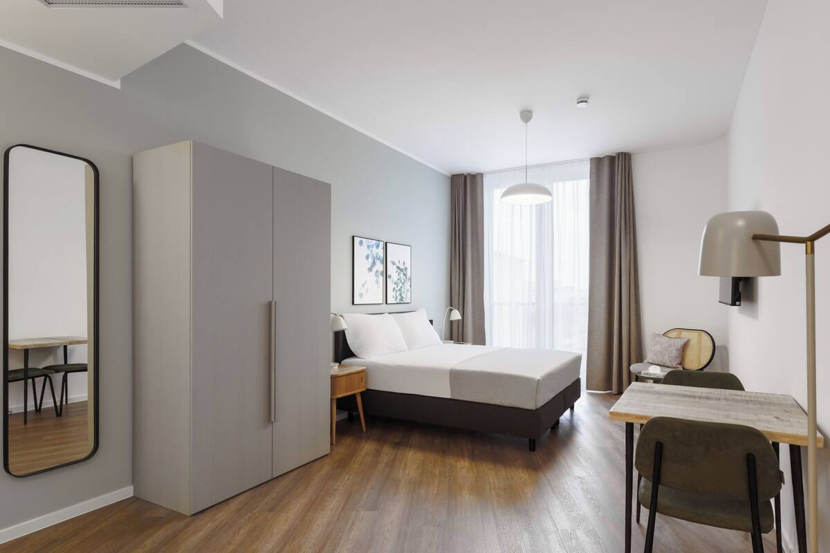 A modern hotel room with a bed, bedside table, lamp, and a desk with a chair.