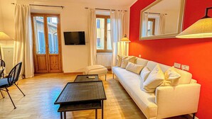 A living room with a white sofa, a black coffee table, a TV mounted on the wall, and a window with curtains. - RED81 Suite - 10 min dal Duomo - 2 camere (Milan)